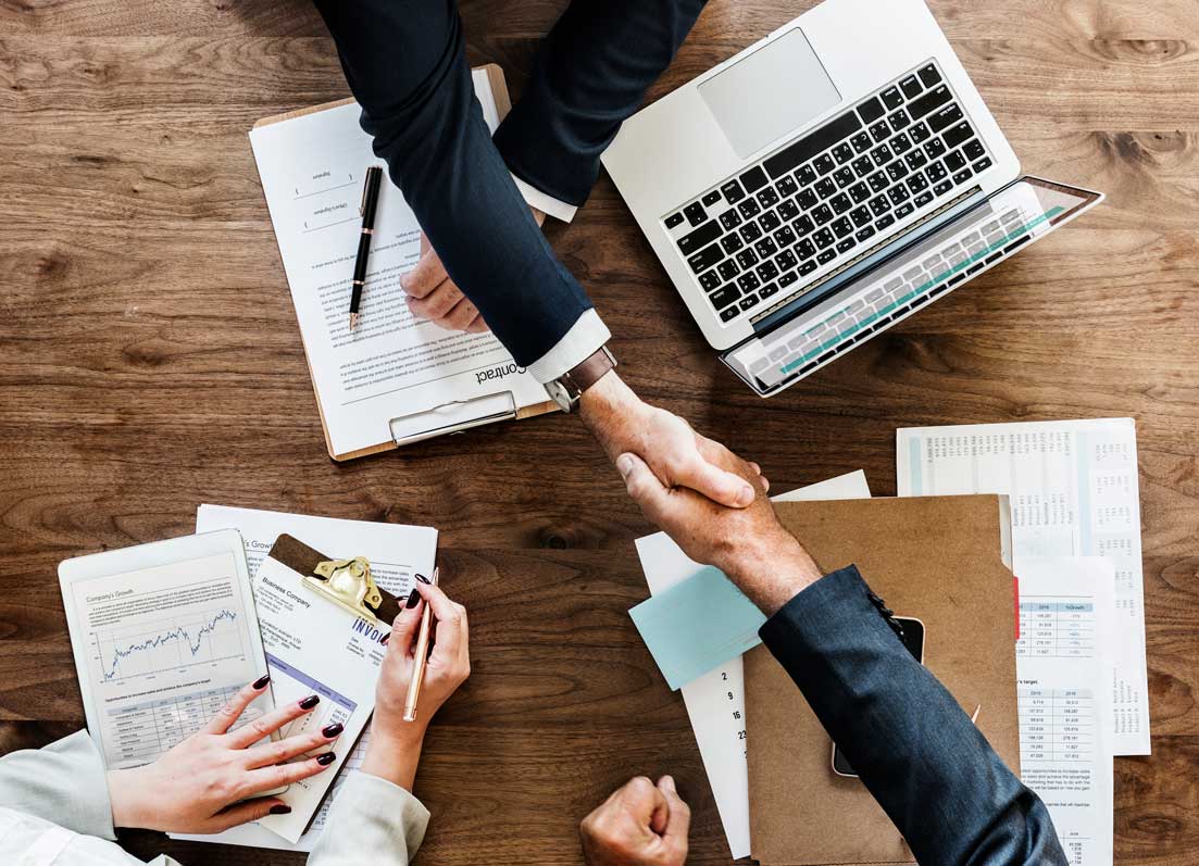 Seen from above, two people shake hands across a desk of invoices, spreadsheets and a laptop.