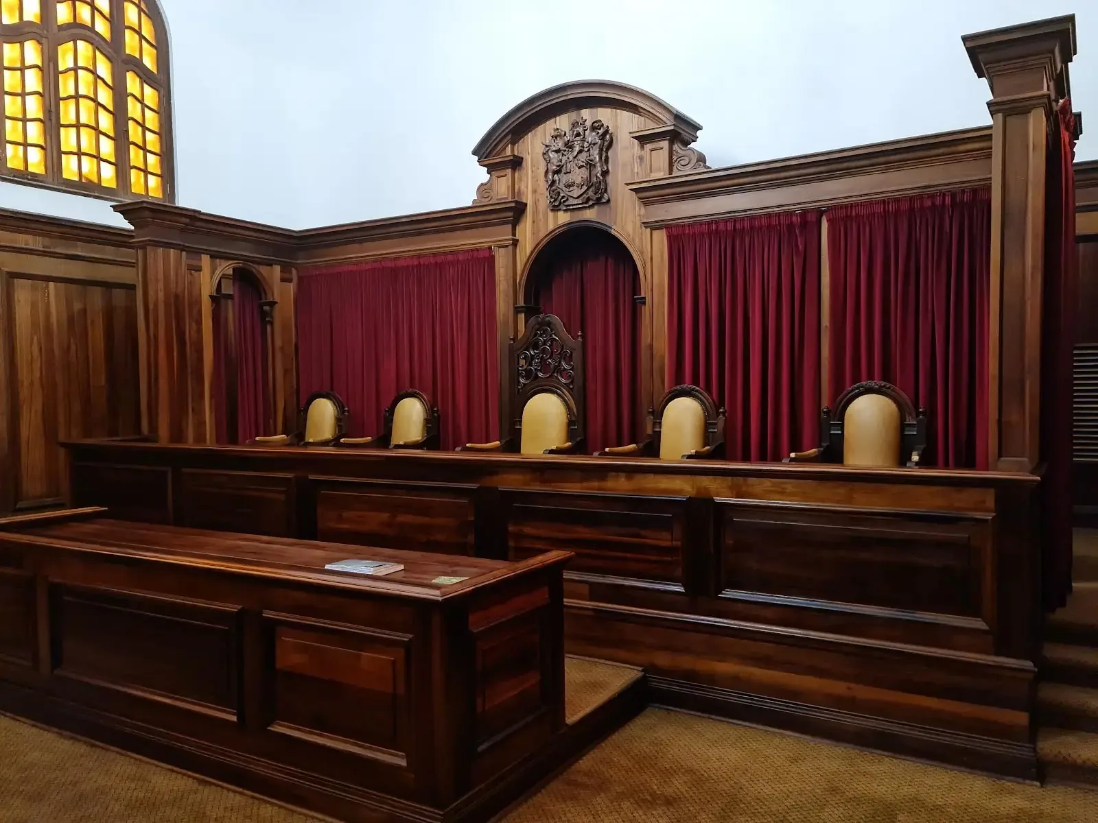 A wood-panelled South African courtroom: five judges’ chairs at the bench beneath a carved coat of arms, red velvet curtains and an amber stained-glass window.